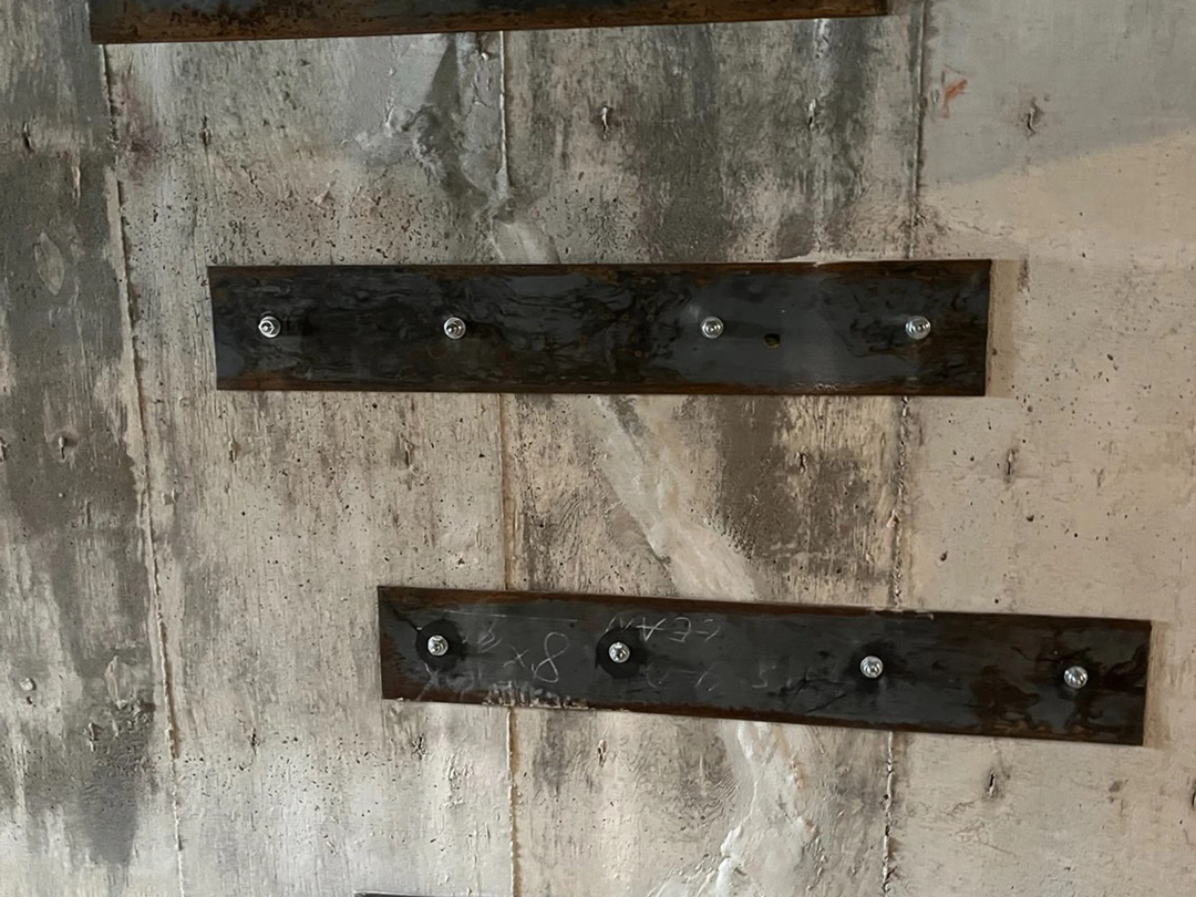 CRACK REPAIR Rusty metal plates affixed to a textured concrete wall, each plate secured with visible screws, showcasing a weathered industrial aesthetic