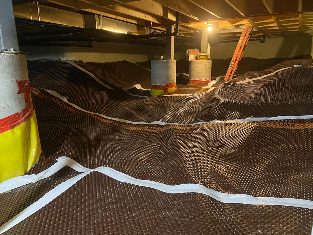 CRAWL SPACE DRAINAGE Underground space with a brown tarp covering uneven ground, yellow and red wrapped posts, and a ladder illuminated by overhead lighting