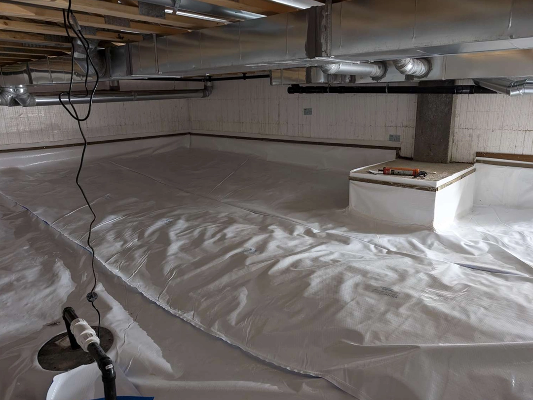 CRAWL SPACE LINER A basement with a white vapor barrier covering the floor, pipes overhead, and a small raised area with construction tools nearby