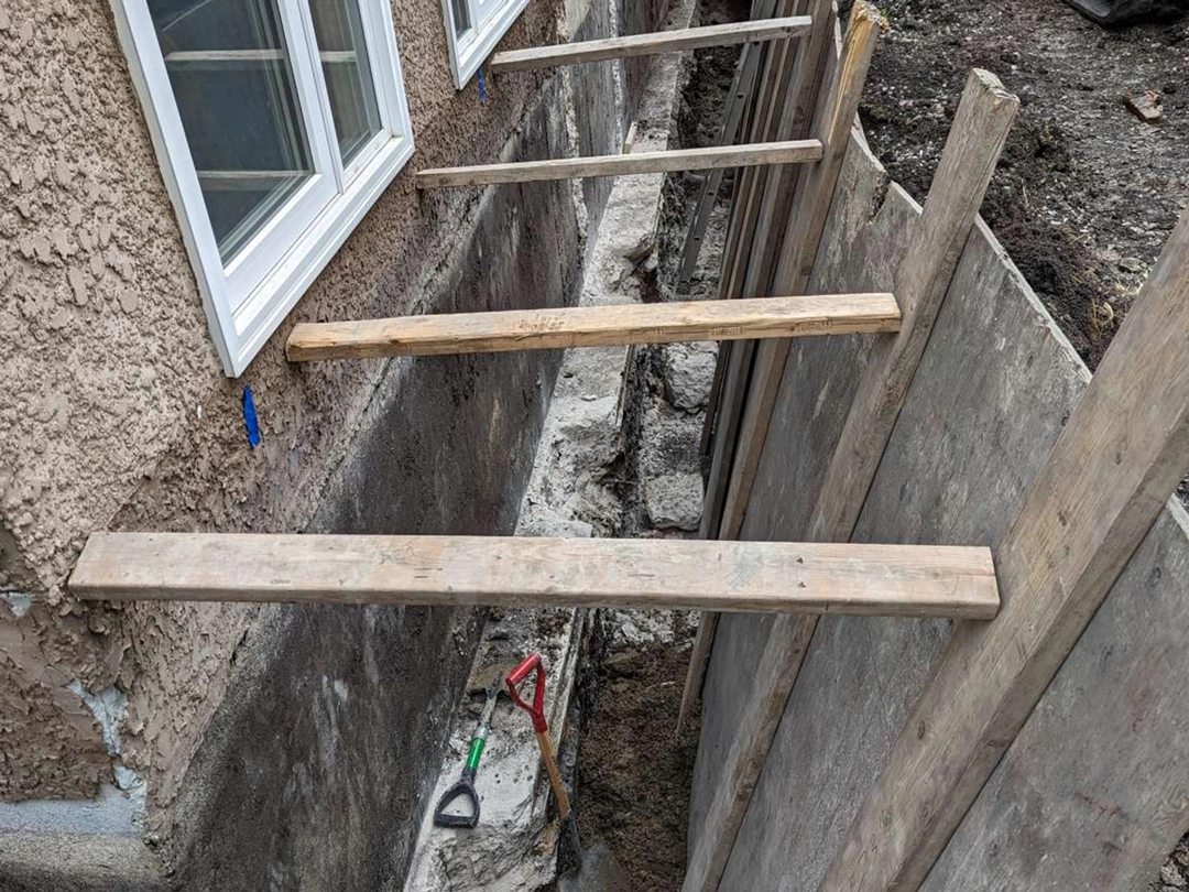 Foundation Underpinning Wooden planks extend over a trench next to a house, with tools visible on the ground and unfinished concrete walls in the background