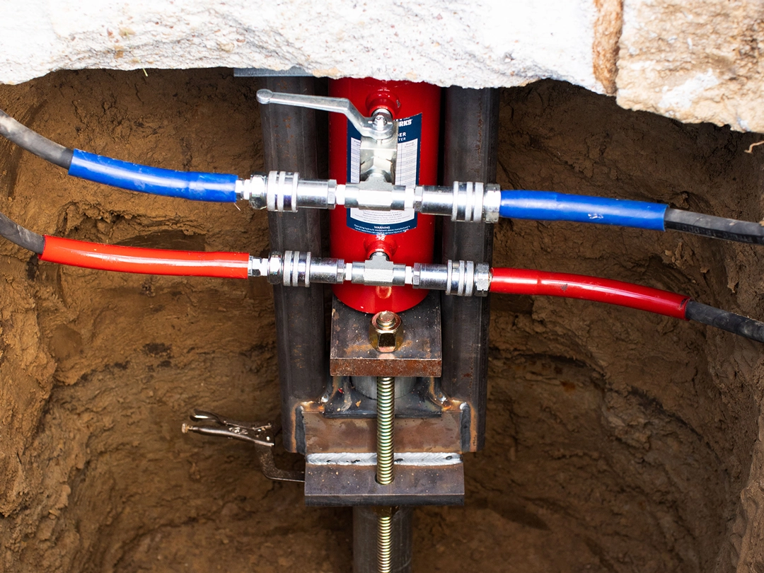 HOUSE AND BUILDING LEVEL Close-up of a red hydraulic pump in a trench, with blue and red hoses connected to it, set in earthy surroundings