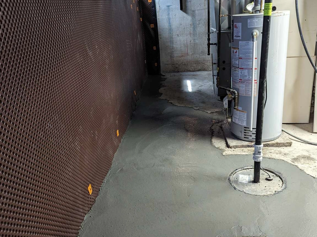 INTERIOR DRAINAGE SYSTEMS Freshly poured concrete floor in a basement with a water heater and drainage pipe nearby, against a textured brown wall