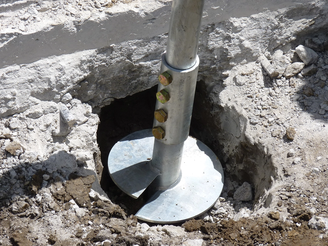STEEL PIER SYSTEM A large metal helical anchor installed in soil, with visible bolts and surrounding excavated earth