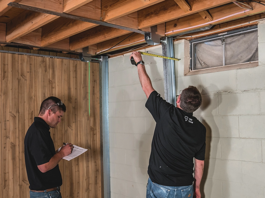 WALL BRACING Two men in black shirts are measuring and inspecting a basement area, with one holding a measuring tape and the other taking notes