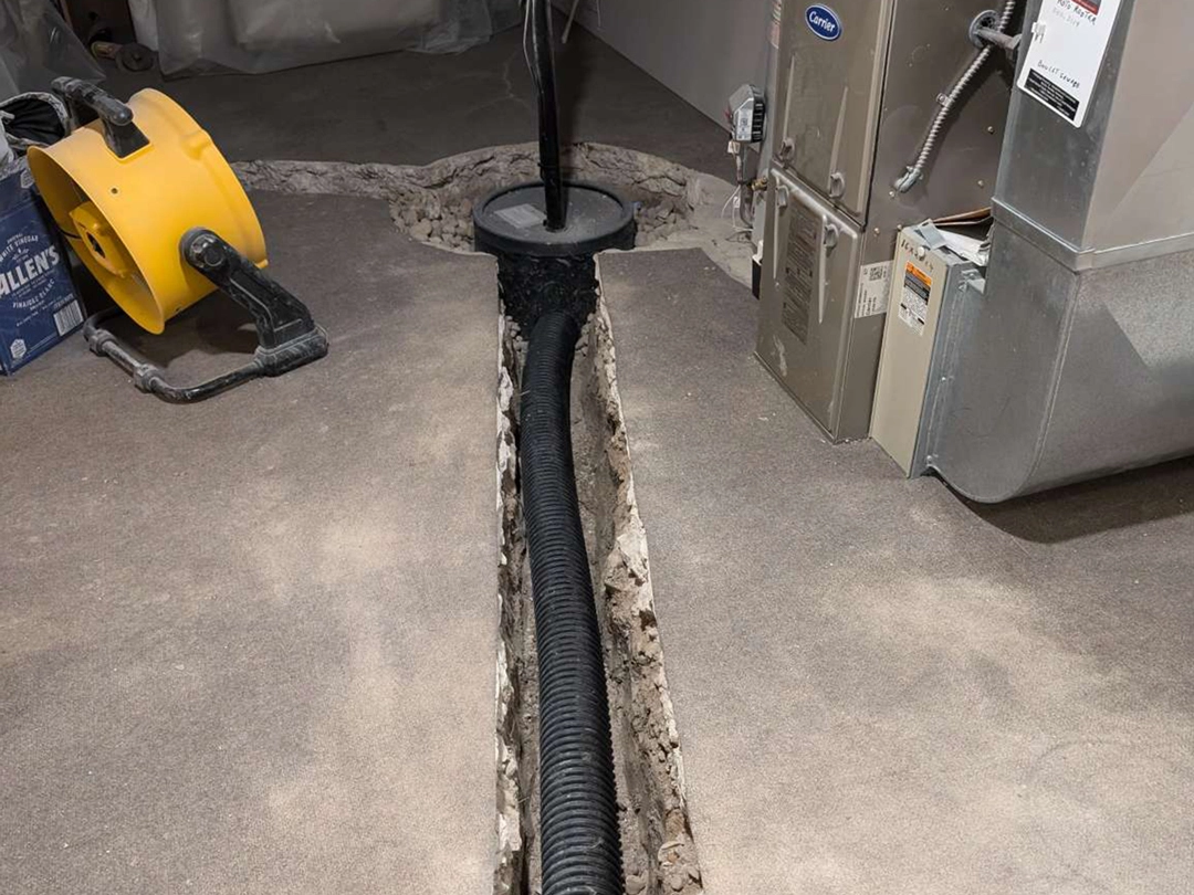 WEEPING TILE A basement with a cutout floor revealing a drainage trench, black piping, and a sump pump, alongside a yellow blower and HVAC unit